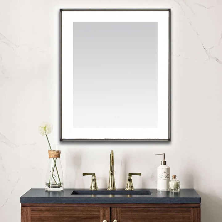 A modern bathroom vanity with a dark sink, gold faucet, and wooden cabinet is topped by the Lexington 30 inch x 36 inch LED Lighted Slim Frame Mirror. A glass vase with a single flower and various accessories rest on the counter.