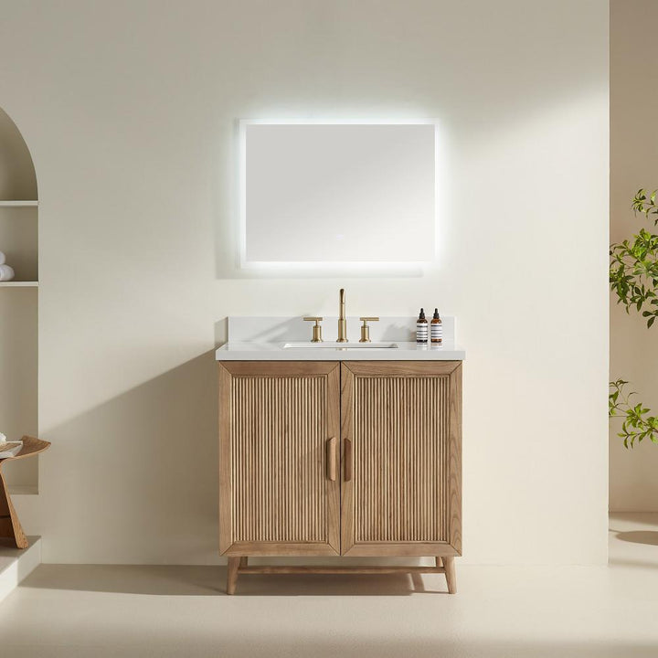 The Liora 30 Inch Bathroom Vanity features a light wood cabinet, white countertop, gold faucet, two small bottles, and a large illuminated rectangular mirror on a neutral wall, with shelves and a plant in the background.