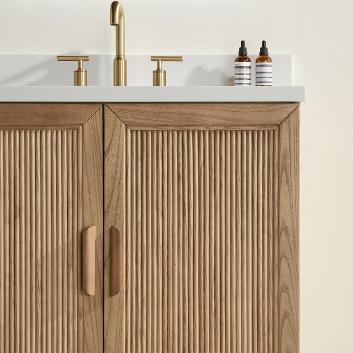 The Liora 30 Inch Bathroom Vanity features vertical wood slat cabinet doors, gold faucet and handles, plus two small bottles with striped labels on a white countertop.