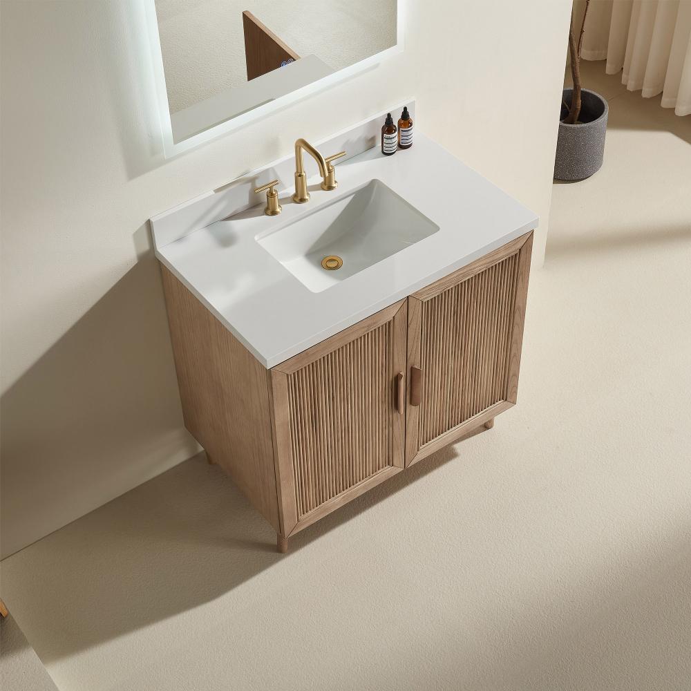 The Liora 30 Inch Bathroom Vanity features a white rectangular sink, gold faucet, and wooden doors. It’s set against a light wall with two small bottles and a mirror above, complemented by neutral-toned floors and curtains.