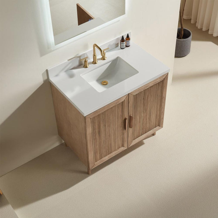 The Liora 30 Inch Bathroom Vanity features a white rectangular sink, gold faucet, and wooden doors. It’s set against a light wall with two small bottles and a mirror above, complemented by neutral-toned floors and curtains.