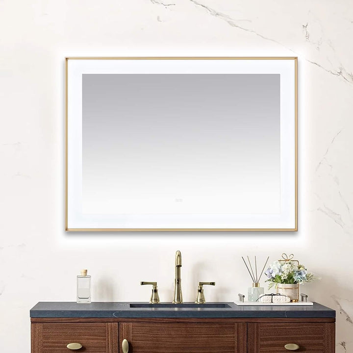 A Lexington 48 inch x 36 inch LED Lighted Slim Frame Mirror with a thin gold frame hangs above a dark wood vanity with black countertop, gold faucet, soap dispenser, and small floral arrangement against a light marble-veined wall.