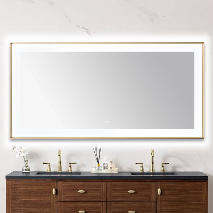 A modern bathroom features a Lexington 72 inch x 36 inch LED Lighted Slim Frame Mirror above a double-sink vanity with brass faucets, wooden cabinets, and minimal decor like a vase with white flowers and a reed diffuser.