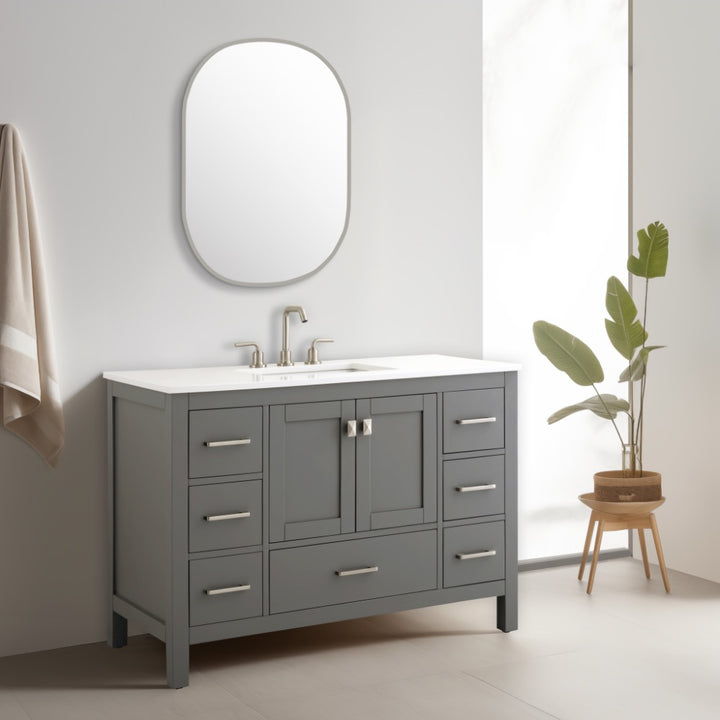 A modern bathroom showcases the Vanea 48 inch Bathroom Vanity in gray with a white quartz top, silver handles, an oval wall mirror, a beige towel, and a green plant in a wooden stand by a frosted window.