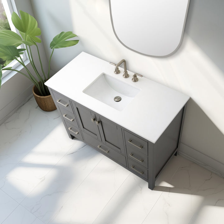 The Vanea 48 inch Bathroom Vanity in Gray with a white quartz top features spacious drawers. Sunlight streams across the white tile floor, highlighting the vanity as a green potted plant sits nearby.