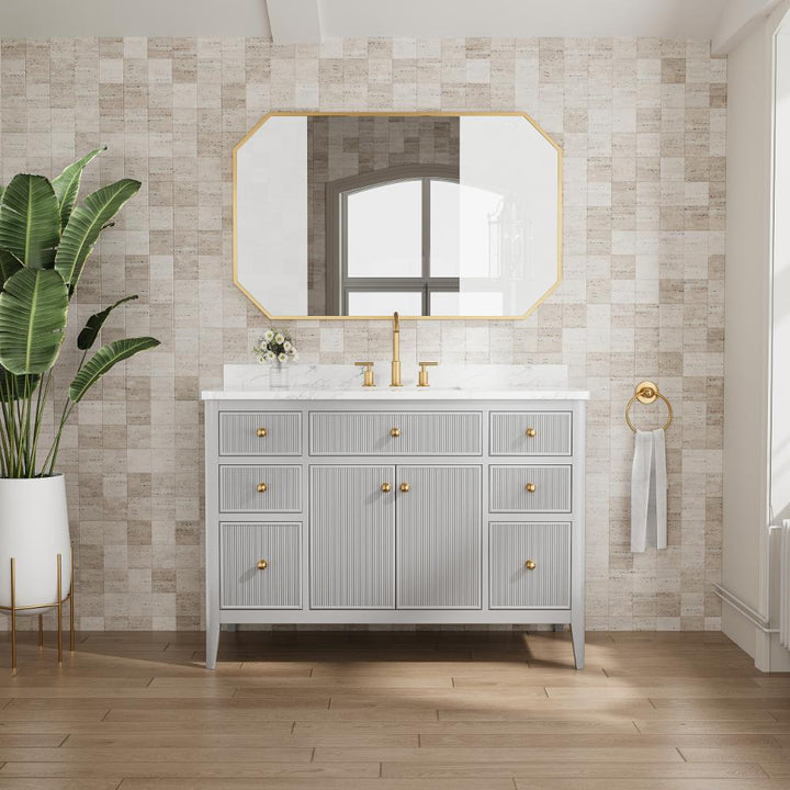 A modern bathroom features the Aveline 48 Inch Freestanding Bathroom Vanity with Top in light gray and gold hardware beneath a large geometric mirror. A potted plant stands to the left, with a towel ring and window on the right.