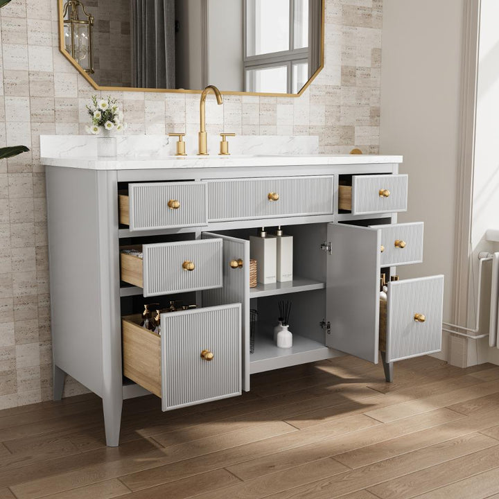 A modern Aveline 48 Inch Freestanding Bathroom Vanity With Top features a white countertop, gold faucet, multiple open drawers and cabinets, light gray finish with gold handles, set against brown floor tiles and a large mirror in the background.
