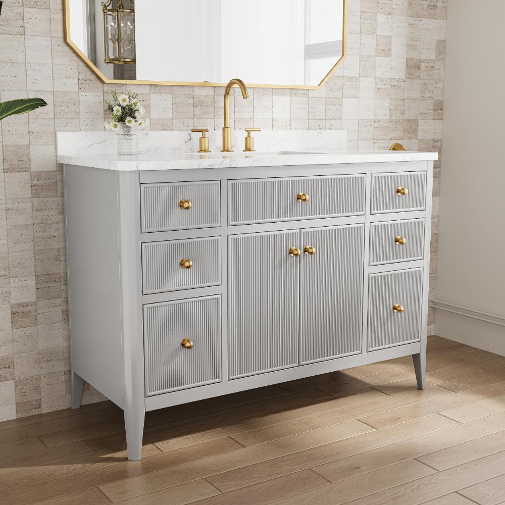 The Aveline 48 Inch Freestanding Bathroom Vanity With Top features a white marble countertop, gold faucet and handles, gray cabinets, a small flower vase under a gold-framed mirror, all set against a textured beige tile wall.