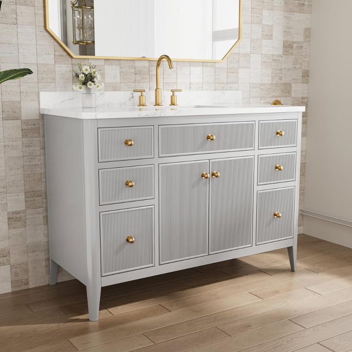 The Aveline 48 Inch Freestanding Bathroom Vanity With Top features a white marble countertop, gold faucet and handles, gray cabinets, a small flower vase under a gold-framed mirror, all set against a textured beige tile wall.