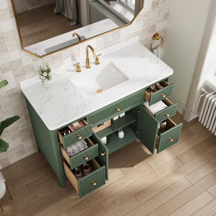 The Aveline 48 Inch Freestanding Bathroom Vanity With Top features a white marble countertop, gold faucet, green cabinets with eight drawers—some open to show storage—and a small flower arrangement next to the sink beneath a mirror on the tiled wall.
