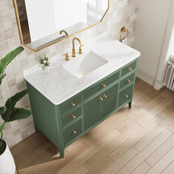 The Aveline 48 Inch Freestanding Bathroom Vanity With Top features a green finish, gold handles, and a white marble countertop. Gold faucet, rectangular sink, mirror, small plant, soap dispenser, and light wood flooring complete the look.