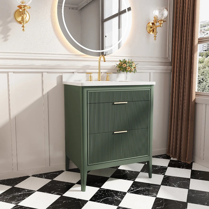 A modern bathroom features the Aveline 30 Inch Freestanding Bathroom Vanity With Top in green with gold hardware, a white countertop, a round backlit mirror, gold wall sconces, black-and-white checkered floor tiles, and a small plant on the counter.