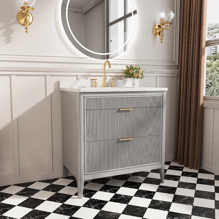 A modern bathroom features the Aveline 36 Inch Freestanding Bathroom Vanity with Top, gray fluted front, gold faucet, round mirror, wall sconces, small flower vase, black-and-white checkered tile floor, white wainscoting, and a window.