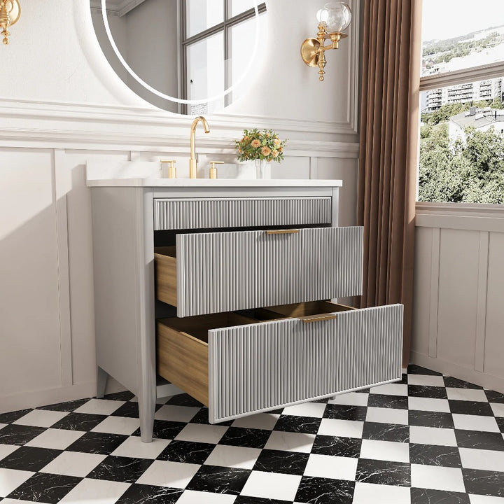 The Aveline 36 Inch Freestanding Bathroom Vanity With Top includes two open drawers, gold faucet, round mirror, small flower vase, wall sconce, and sits on a black-and-white checkered floor beside a window with brown curtains showing a city view.