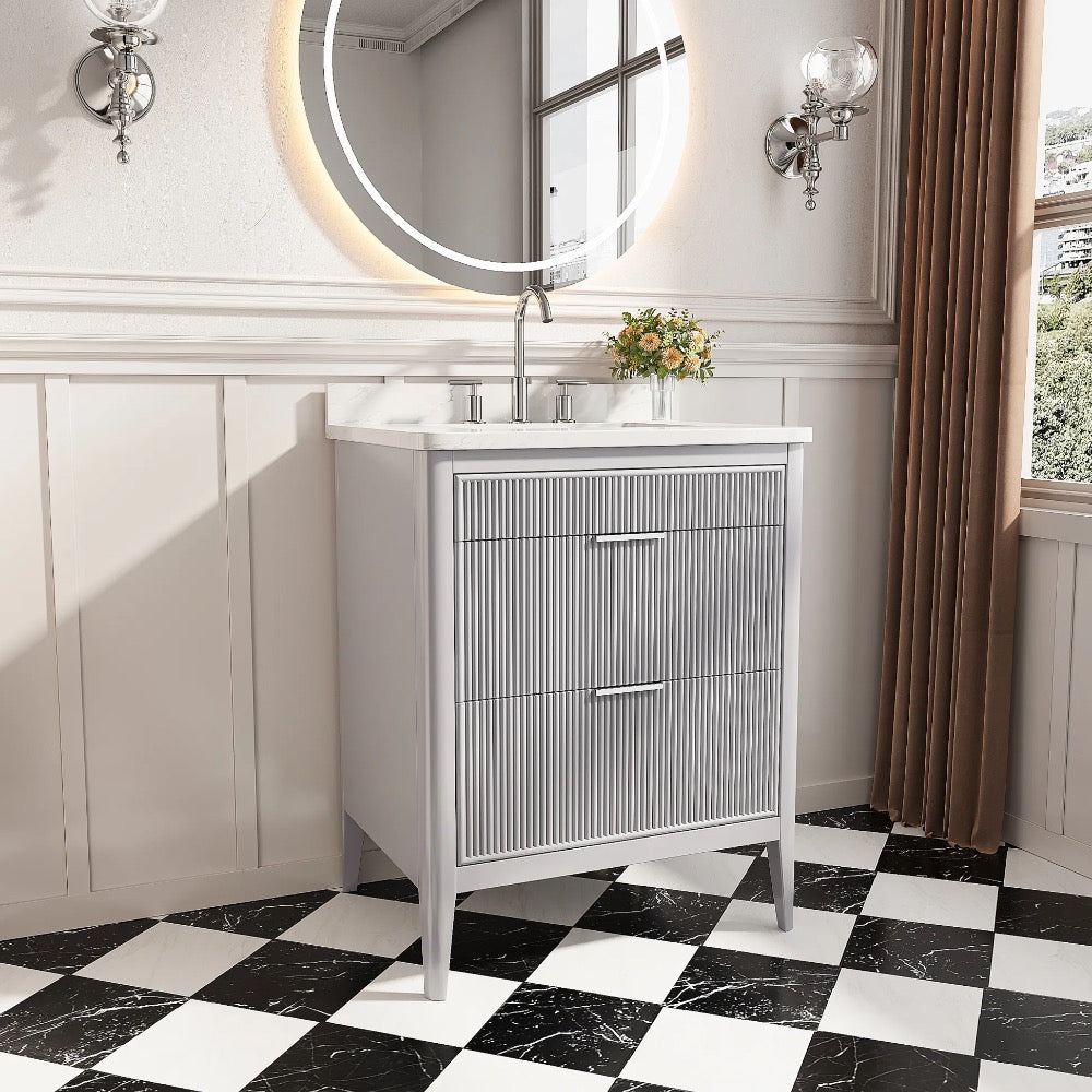 The Aveline 30 Inch Freestanding Bathroom Vanity With Top features a ribbed gray cabinet, white countertop, faucet, round backlit mirror, wall sconces, small vase of flowers, and a black-and-white checkered tile floor.