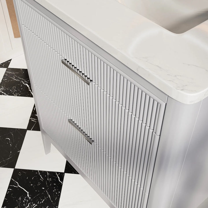 A close-up of the Aveline 30 Inch Freestanding Bathroom Vanity With Top highlights vertical grooves on the drawers, metal handles, and a white marble countertop. The floor showcases a black and white checkered tile pattern.