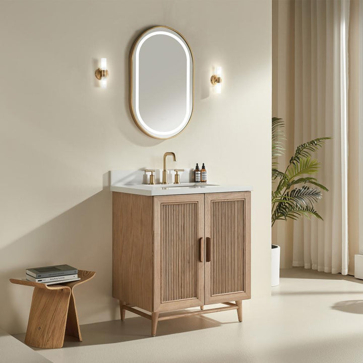 A light wood Liora 36 Inch Bathroom Vanity with a white countertop, gold faucet, and bottles sits below an oval gold-framed mirror. Modern decor is completed by wall sconces, a plant, beige walls, curtains, and a small stool with books.