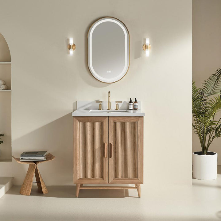 Minimalist bathroom featuring a Liora 36 Inch Bathroom Vanity with white countertop, oval mirror, gold fixtures, twin wall sconces, a potted plant, and a small wooden stool with books—all set against a beige wall.