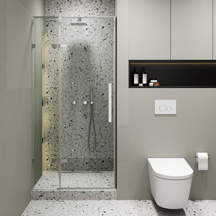 Modern bathroom with a Nova Hinged Frameless Shower Door enclosing terrazzo walls and floor, wall-mounted toilet, toilet paper holder, recessed shelf with toiletries, and neutral-toned walls and cabinetry.