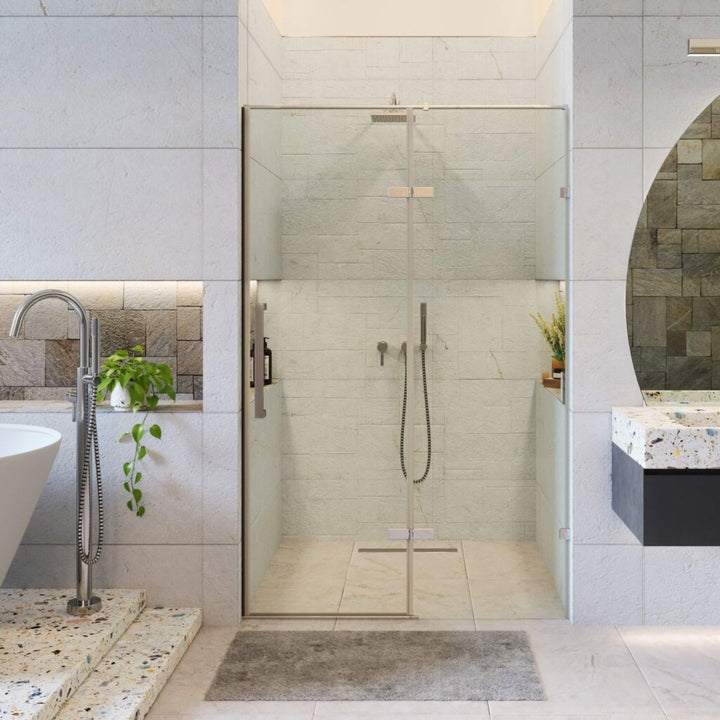 A modern bathroom features the Nova Hinged Frameless Shower Door, a white freestanding bathtub, terrazzo sink, wall plants, grey stone walls and flooring, and a gray mat in front of the shower.