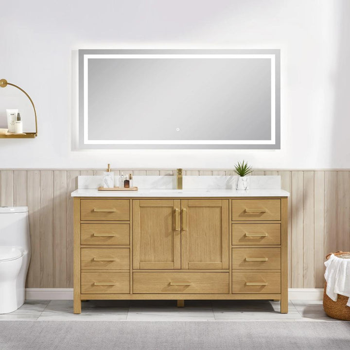 A modern bathroom showcases the Basel 60 Inch Single Bathroom Vanity with gold hardware, a white countertop, and dual sinks. A large lighted mirror hangs above; to the left, a toilet sits beneath a wooden shelf holding toiletries and a plant.