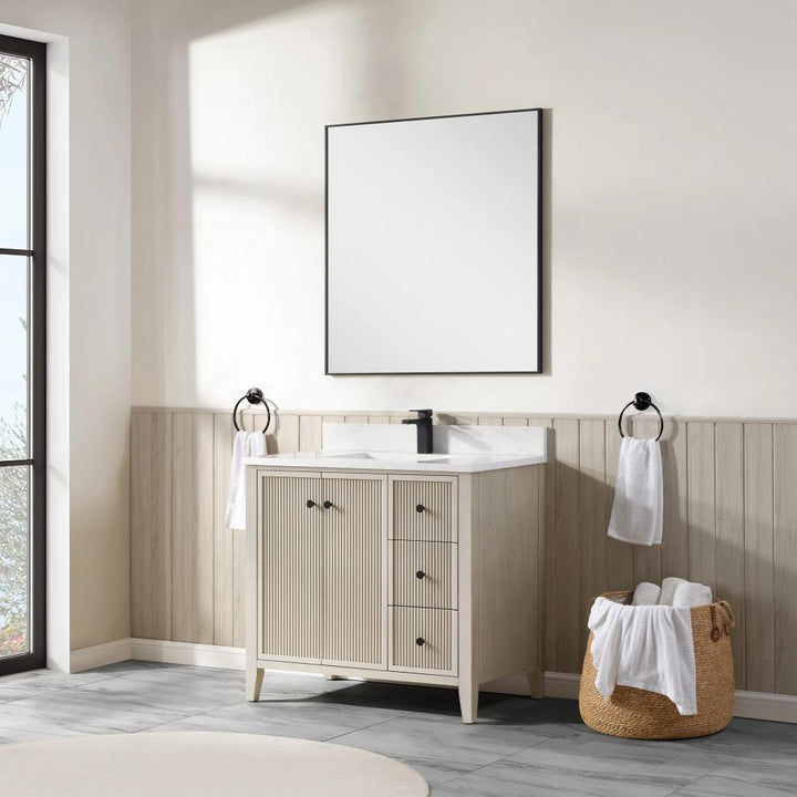 A modern bathroom features the Torino 36 Inch Bathroom Vanity Cabinet with drawers, black faucet, square mirror, towel rings with white towels, and a woven basket on a gray tile floor. Sunlight streams in from a large window.