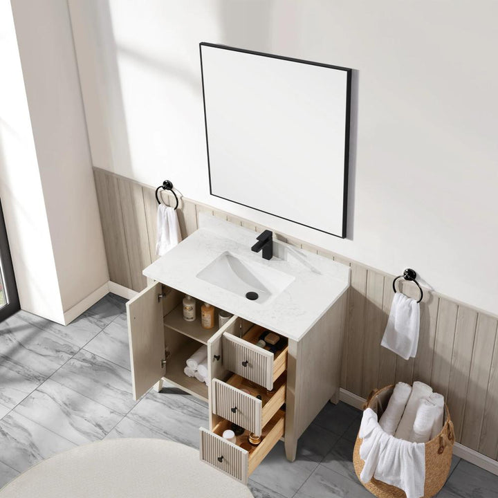 A modern bathroom featuring the Torino 36 Inch Bathroom Vanity Cabinet with drawers, open for display, paired with a black faucet, a square mirror, towel rings holding white towels, and a basket of rolled towels on a gray marble floor.