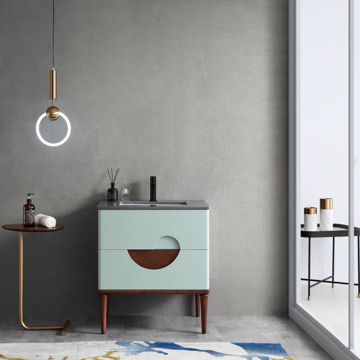 Modern bathroom with a sleek circular pendant light, VABURAG30FD 30-inch Aloe Green Freestanding Bathroom Vanity with a semicircular cutout, and a gold side table holding a soap dispenser and towel. A glass door reveals large containers outside, and a blue and white rug adorns the floor.