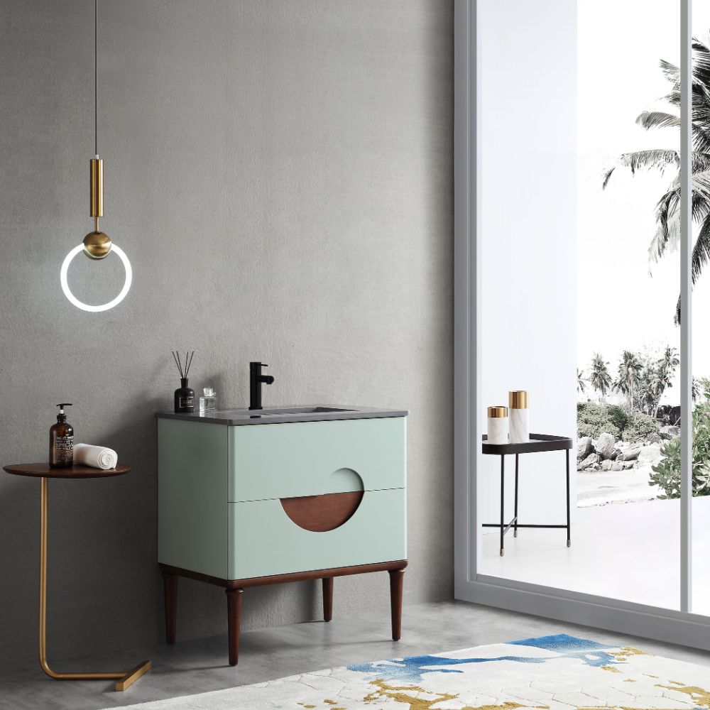 A modern bathroom features the Burano 30 inch Aloe Green Freestanding Bathroom Vanity with wooden legs, a round wall light, a small side table holding soap, and a large window displaying tropical palm trees outside.