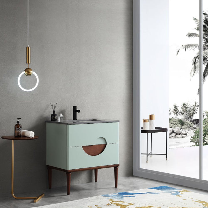 A modern bathroom features the Burano 30 inch Aloe Green Freestanding Bathroom Vanity with wooden legs, a round wall light, a small side table holding soap, and a large window displaying tropical palm trees outside.