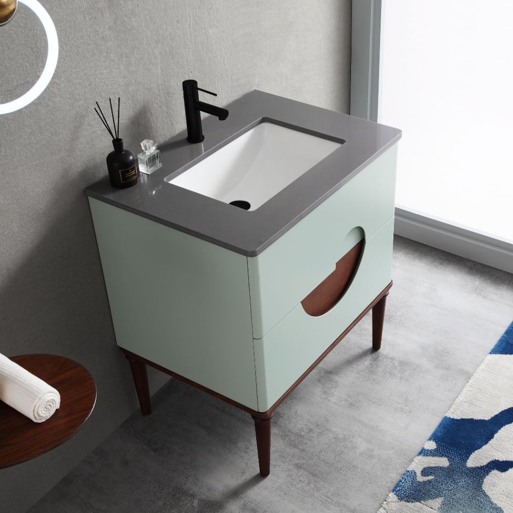 The Burano 30 inch Aloe Green Freestanding Bathroom Vanity features a light green cabinet with a circular cutout, gray countertop, rectangular white sink, black faucet, and is complemented by decorative items and a blue patterned rug.