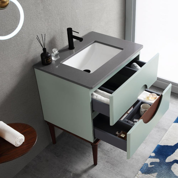The Burano 30 inch Aloe Green Freestanding Bathroom Vanity has a gray countertop, white sink, and black faucet, with two open drawers holding toiletries. A towel, round mirror, and patterned rug are also partially visible.
