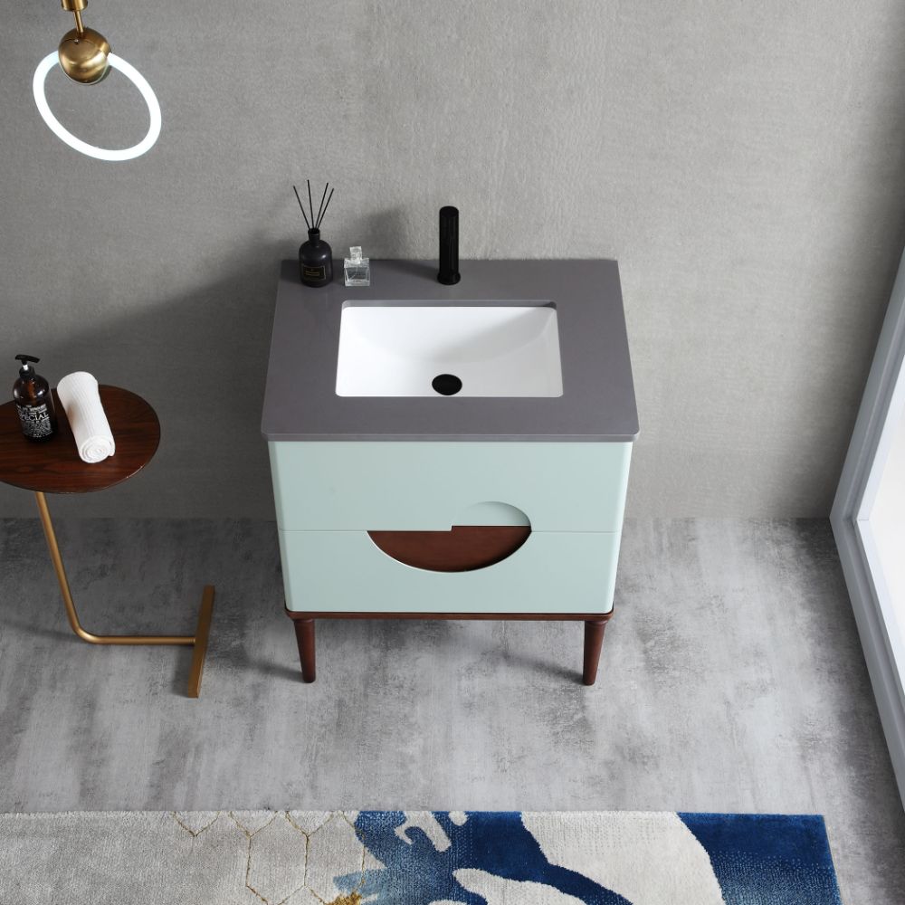 The Burano 30 inch Aloe Green Freestanding Bathroom Vanity has a square sink, gray countertop, and light blue cabinet with a circular cutout. It stands on wooden legs, with a small side table holding toiletries and a towel nearby.