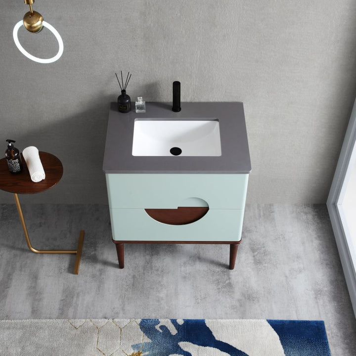 The Burano 30 inch Aloe Green Freestanding Bathroom Vanity has a square sink, gray countertop, and light blue cabinet with a circular cutout. It stands on wooden legs, with a small side table holding toiletries and a towel nearby.