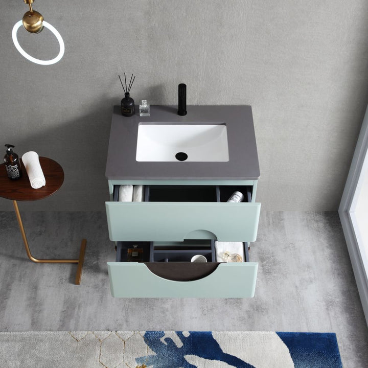The Evano 30 inch Aloe Green Freestanding Bathroom Vanity features a grey countertop, white sink, black faucet, and two open drawers for towels and toiletries. A small side table with more toiletries sits on a grey floor with a blue patterned rug.