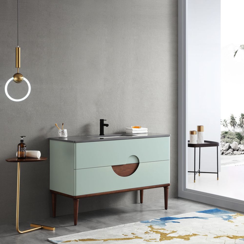 A modern bathroom features the Burano 48 inch Aloe Green Freestanding Bathroom Vanity with wooden legs and accents, a black faucet, a round side table, pendant light, and a large window with an outdoor view.