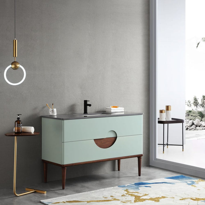 A modern bathroom features the Burano 48 inch Aloe Green Freestanding Bathroom Vanity with wooden legs and accents, a black faucet, a round side table, pendant light, and a large window with an outdoor view.