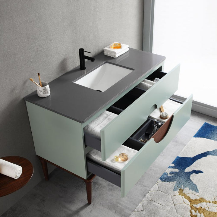 The Burano 48 inch Aloe Green Freestanding Bathroom Vanity features a gray countertop, black faucet, built-in sink, and two open drawers with towels and toiletries. A round side table with a rolled towel sits on a patterned rug nearby.