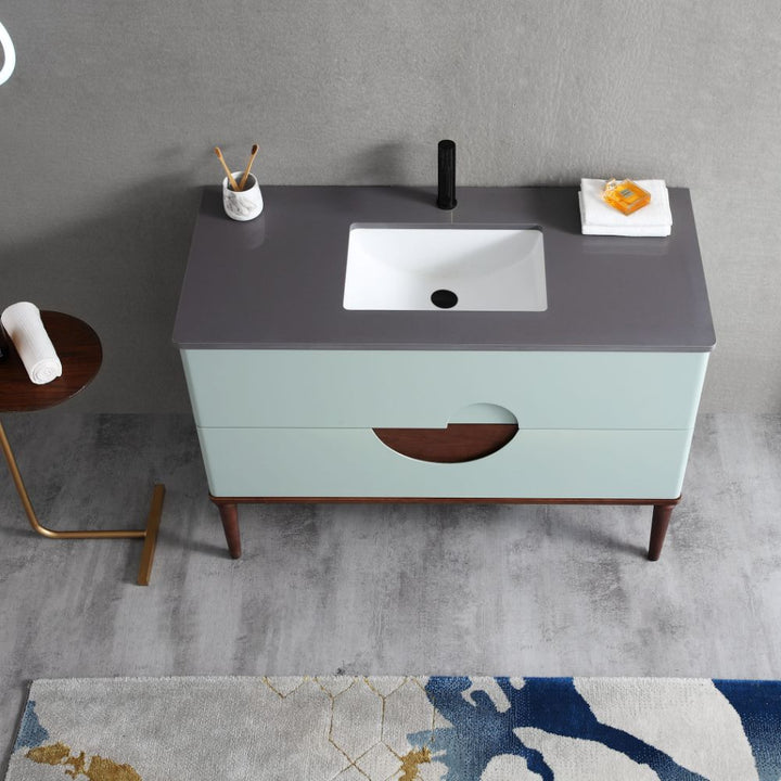 The Burano 48 inch Aloe Green Freestanding Bathroom Vanity features a gray countertop, white rectangular sink, black faucet, and two drawers. It’s styled with a soap dish, towels, a toothbrush holder on top, set against a gray wall with a patterned rug.
