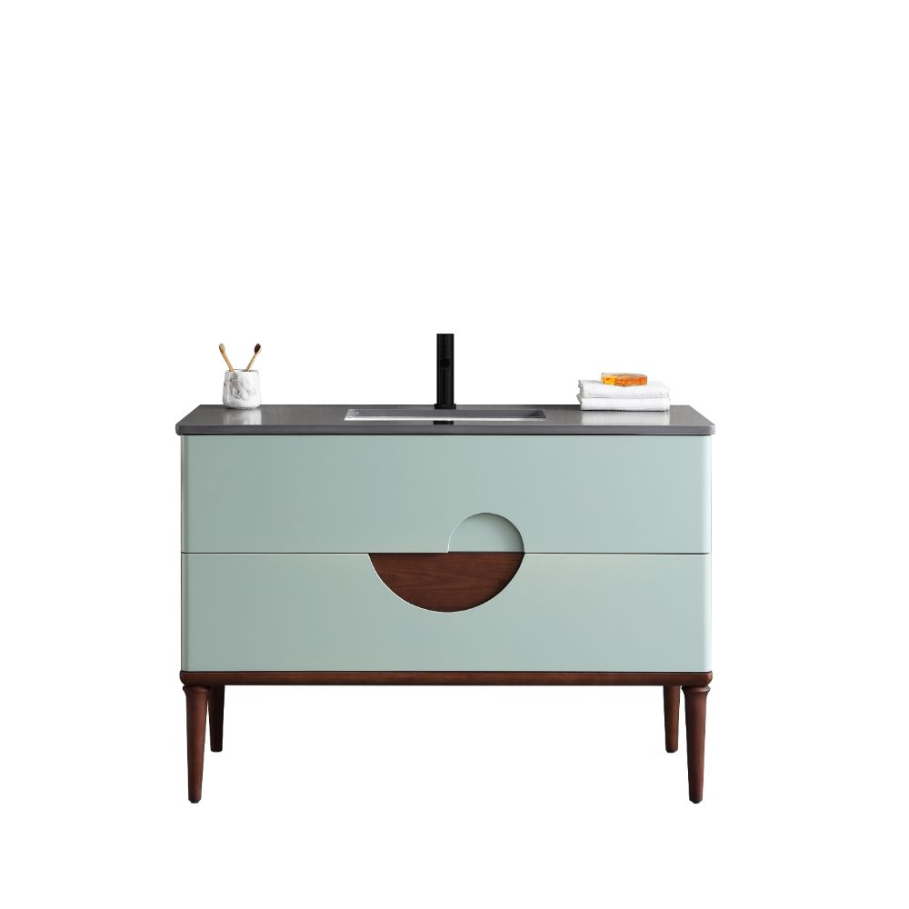 The Burano 48 inch Aloe Green Freestanding Bathroom Vanity features light blue drawers, a circular wood accent, black faucet, dark countertop, and tall wooden legs. A toothbrush holder sits left; towels are stacked right.