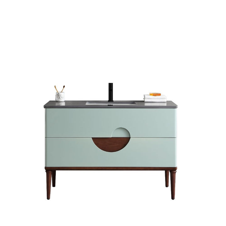 The Burano 48 inch Aloe Green Freestanding Bathroom Vanity features light blue drawers, a circular wood accent, black faucet, dark countertop, and tall wooden legs. A toothbrush holder sits left; towels are stacked right.