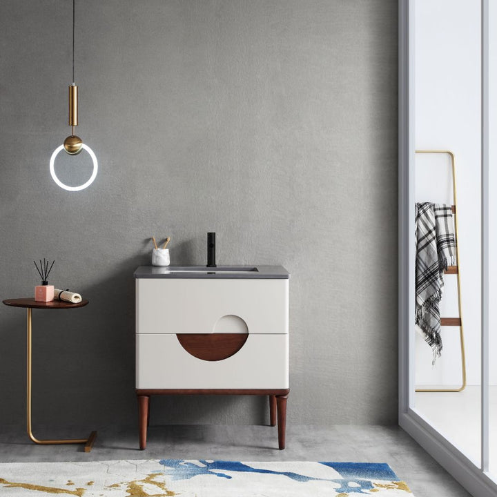 A minimalist bathroom showcases a gray wall and the Burano 30 inch Beige Freestanding Bathroom Vanity with circular wooden detail. A ring-shaped pendant light illuminates a small round table holding a book and brush. Nearby, an abstract-patterned rug enhances the floor beside a towel rack by the glass door.