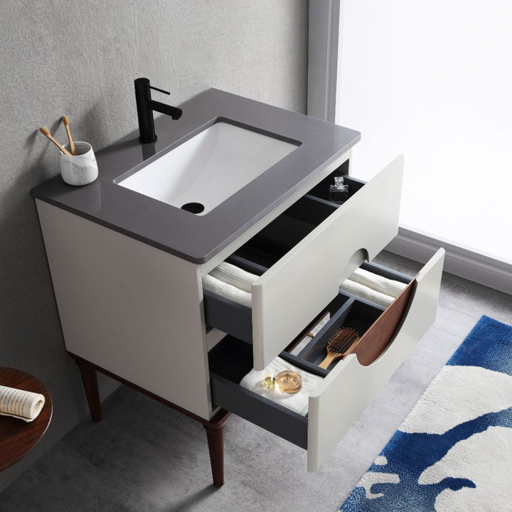 The Burano 30 inch Beige Freestanding Bathroom Vanity features a gray countertop, white sink, black faucet, and two open drawers with toiletries. A toothbrush holder and cup sit atop the vanity, complemented by a blue and white rug on the floor.