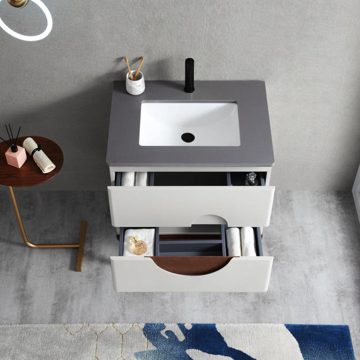 The Burano 30 inch Beige Freestanding Bathroom Vanity has a gray countertop, white sink, and two open drawers with towels and toiletries. Nearby, a side table holds rolled towels and sticks on a gray floor with a colorful rug.