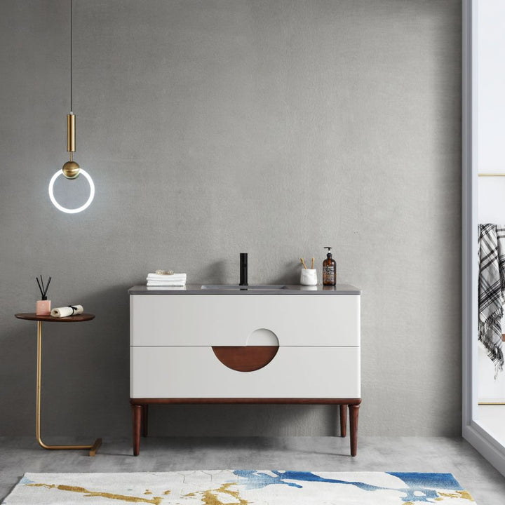 In the minimalist bathroom, the Evano 48-inch Beige Freestanding Vanity with a modern round cutout handle is paired with a sleek black faucet and soap dispenser. A round side table holds decor items on the left, while a pendant light illuminates from above and a textured rug adds warmth below.