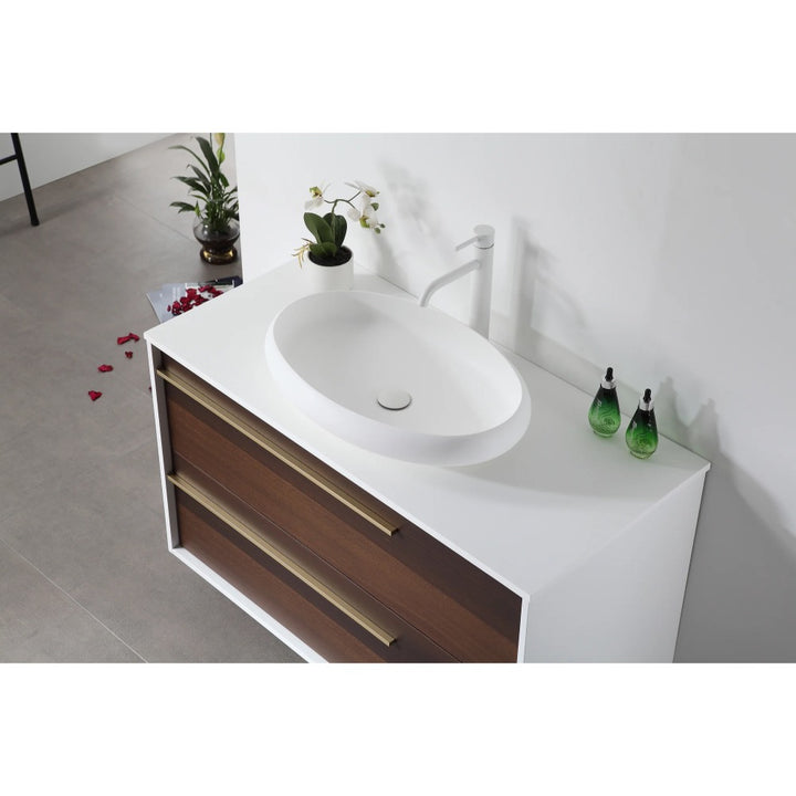 The Goreme 42 Inch Wall Mount Bathroom Vanity With Vessel Sink features a white oval sink, two green glass soap dispensers, and a potted plant on the white countertop. Wooden drawers with gold handles; rose petals scattered on the gray floor.