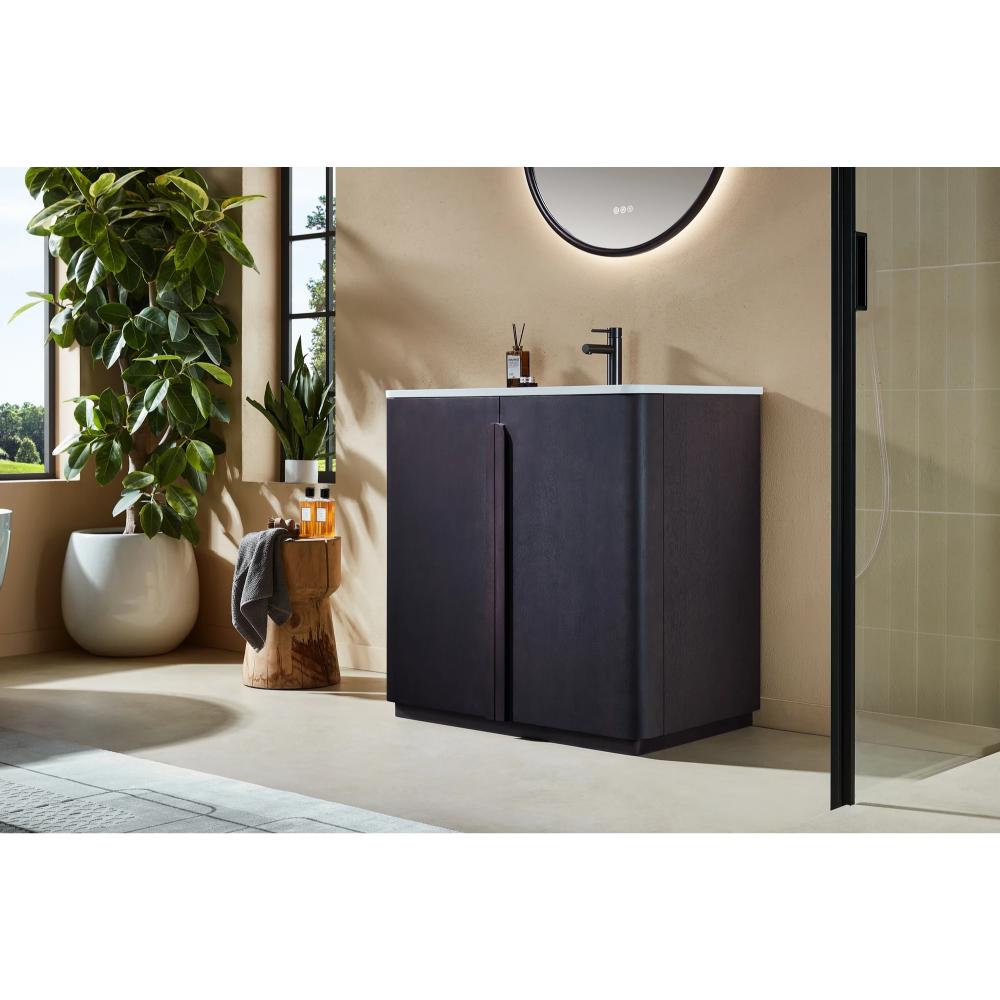 A modern bathroom with the Parma 36 Inch Bathroom Vanity With Top and white sink, a round mirror above, large green plant, sunlit windows, and a wooden stool with bath essentials beside the vanity.