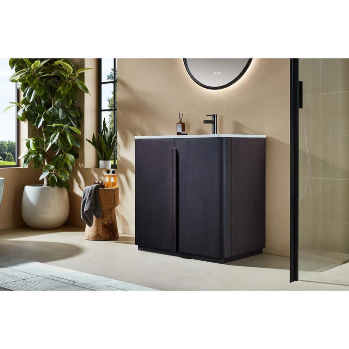 A modern bathroom with the Parma 36 Inch Bathroom Vanity With Top and white sink, a round mirror above, large green plant, sunlit windows, and a wooden stool with bath essentials beside the vanity.