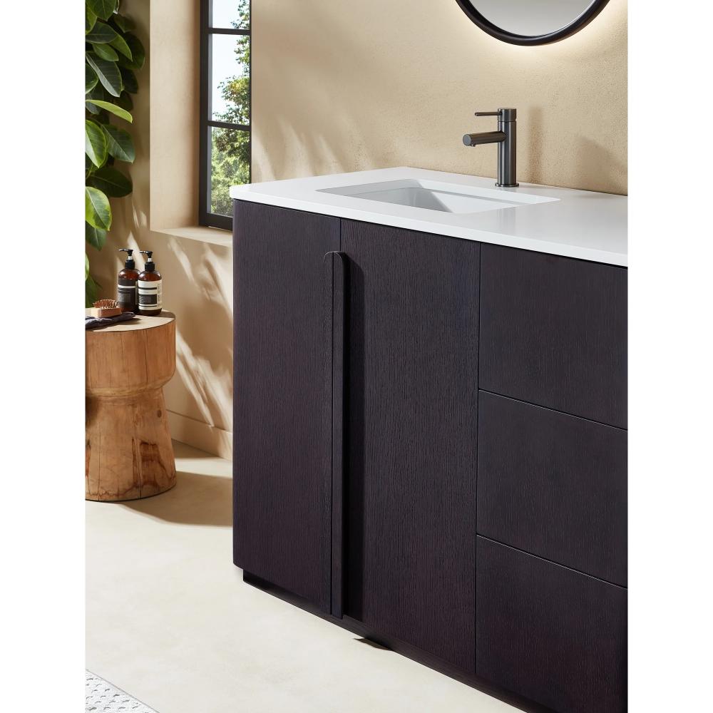 A modern bathroom with the Parma 48 Inch Bathroom Vanity With Top, featuring a dark wood finish, white countertop, and sleek faucet. A round mirror hangs above while a wooden stool with toiletries sits by a leafy plant near the window.