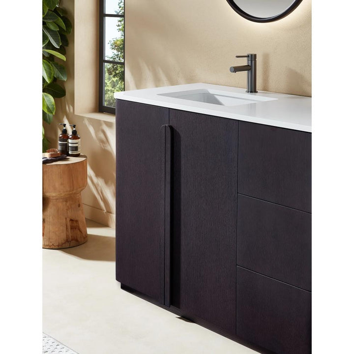 A modern bathroom with the Parma 48 Inch Bathroom Vanity With Top, featuring a dark wood finish, white countertop, and sleek faucet. A round mirror hangs above while a wooden stool with toiletries sits by a leafy plant near the window.